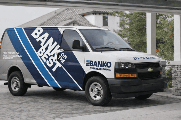 A white Banko Overhead Doors van with blue and black branding is parked in a driveway near a house and garage.