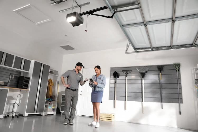 A man in a uniform and a woman stand in a clean, organized garage, looking at a device. Garden tools hang on the wall, and storage cabinets are visible.