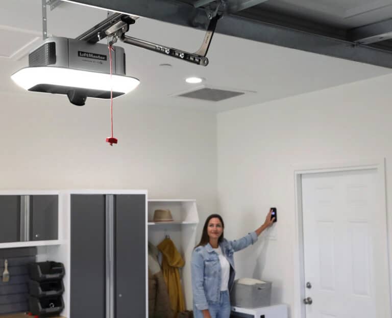 A person stands in a garage holding a remote, with a LiftMaster garage door opener and storage cabinets visible in the background.