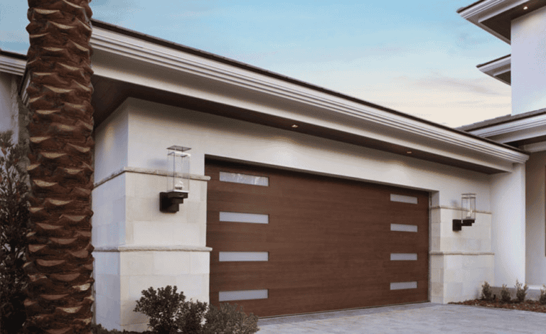 A modern house exterior with a wooden garage door featuring horizontal glass panels, white walls, wall-mounted lights, a palm tree, and small shrubs.