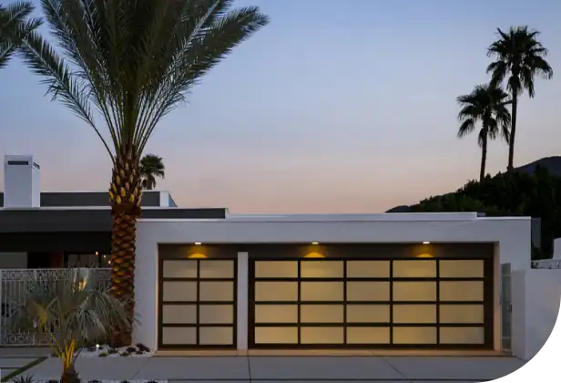 Modern single-story house with a flat roof, large frosted glass garage doors, exterior lighting, and palm trees in the front yard at dusk.