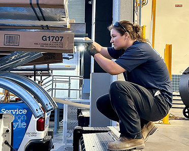 A woman in work clothes crouches on a loading dock, inspecting or organizing stacked boxes and metal parts in an industrial warehouse setting.
