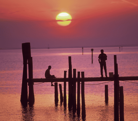 Two people on a wooden dock over calm water at sunset, with the sun low on the horizon and the sky glowing orange and purple.