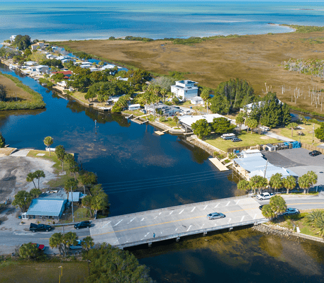 Aerial view of a small canal-side community with houses, trees, a bridge, and cars, bordered by wetlands and the ocean in the background.