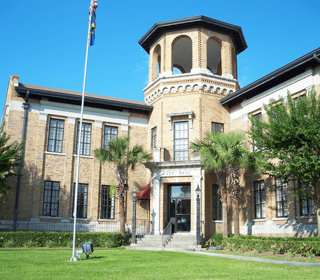 A brick city hall building with an octagonal tower, a flagpole in front, palm trees, and a well-kept lawn under a clear blue sky.