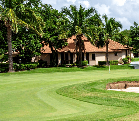 A clubhouse with a brown roof sits behind palm trees and a well-maintained golf course with a putting green and sand trap in the foreground.