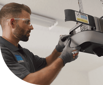 A man wearing safety glasses and gloves installs or repairs a garage door opener in a well-lit indoor space.