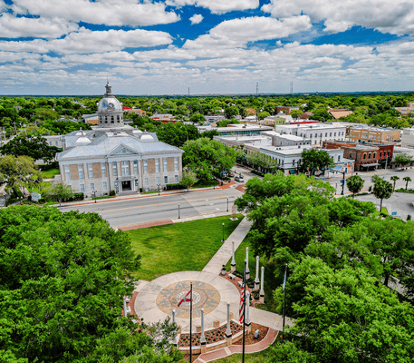 Aerial view of a small town square with a courthouse, surrounding buildings, a circular plaza with flags, and green trees under a partly cloudy sky.