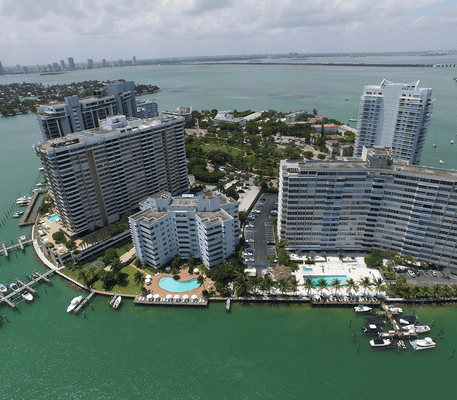 Aerial view of waterfront high-rise buildings, swimming pools, docks, and boats along the edge of a green bay with a distant city skyline.