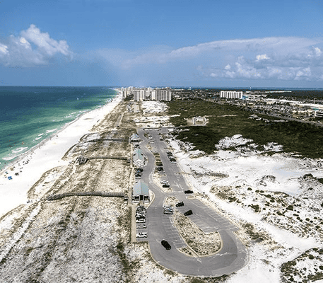 Aerial view of a coastal parking lot near white sand dunes, green vegetation, and high-rise buildings along the shoreline.