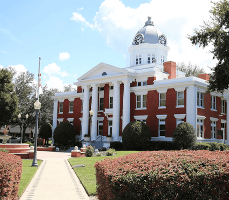 Red brick courthouse with white columns and a clock tower, surrounded by green bushes and trees under a partly cloudy sky.