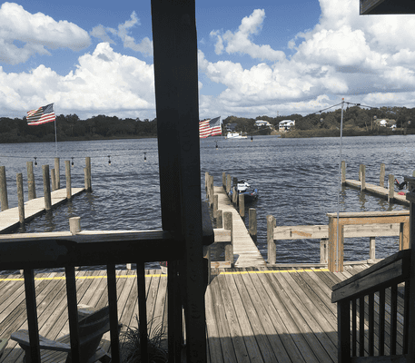 View of a lakeside dock with two American flags, wooden piers, boats, and cloudy sky, seen from a shaded porch area.