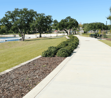 A paved walkway curves through a park with green grass, trimmed bushes, large trees, benches, and a body of water under a clear blue sky.