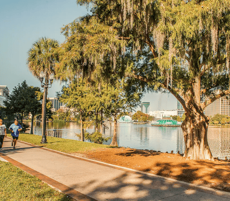 Two people walk on a lakeside path in a city park with trees, a lamppost, and buildings visible in the background on a sunny day.