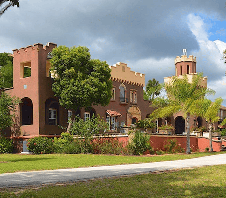 Large stucco building with castle-like towers, arched windows, and palm trees in front, under a cloudy sky.