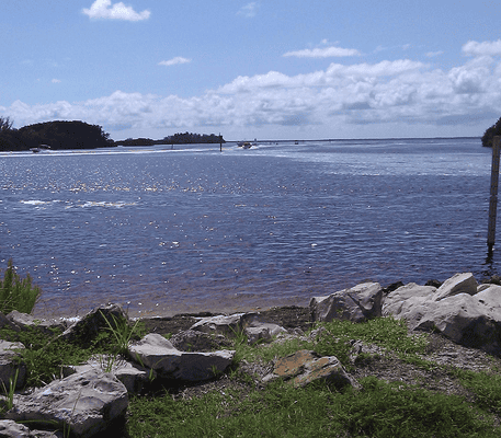 View of a calm body of water with rocks and grass in the foreground, distant boats on the water, and an island and cloudy sky in the background.