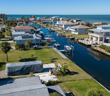 Aerial view of coastal homes lining a narrow canal with docks, boats, and palm trees, leading to the ocean under a clear blue sky.