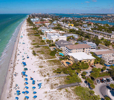 Aerial view of a sandy beach lined with blue umbrellas, next to a coastal town with houses and buildings along the shoreline.