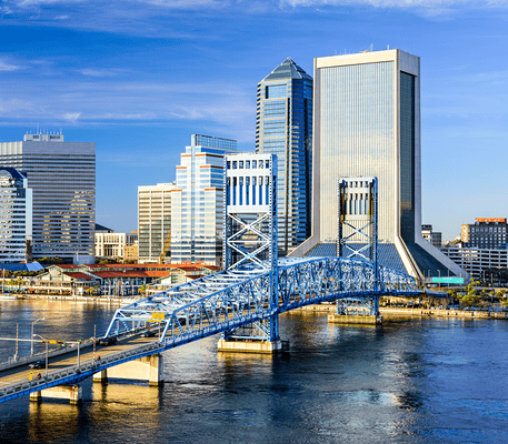 Downtown Jacksonville, Florida skyline with tall buildings and the blue Main Street Bridge over the St. Johns River under a clear sky.