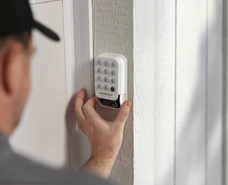A person in a cap is mounting a LiftMaster wireless keypad on a white wooden exterior wall next to a doorframe.