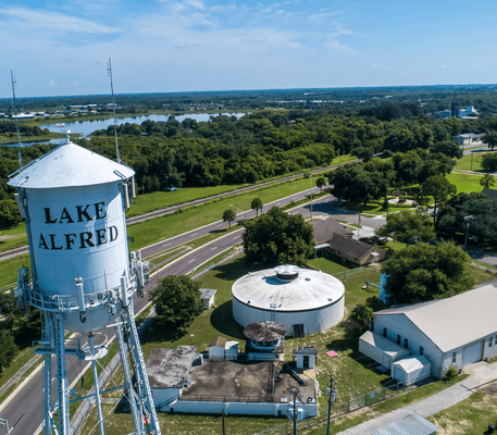 Aerial view of a water tower labeled “Lake Alfred” beside several buildings, roads, and trees with a lake and sky in the background.