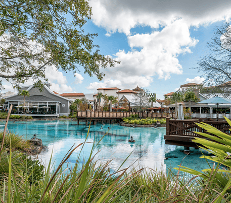 A clear blue pond with wooden walkways, surrounded by landscaping, trees, and several buildings under a partly cloudy sky.