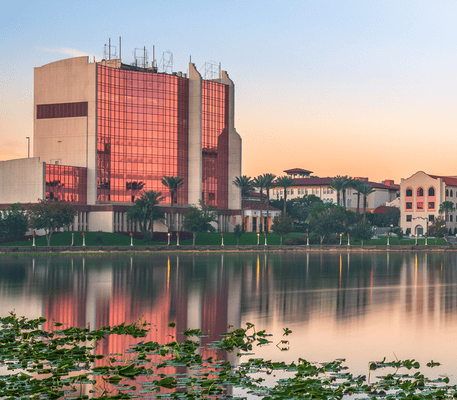 A modern glass building and other structures are reflected in a still lake at sunset, with palm trees and lily pads visible in the foreground.