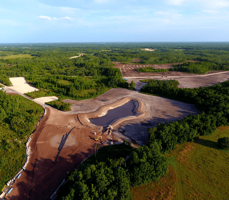 Aerial view of a construction site with cleared land, dirt roads, a pond, and surrounding forest and grassy areas under a blue sky.