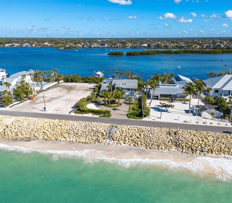 Aerial view of beachfront houses bordered by a stone seawall, clear turquoise water, and blue sky with scattered clouds.