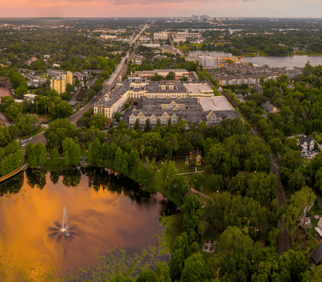 Aerial view of a neighborhood with a pond and fountain, surrounded by trees and buildings, under a colorful sunset sky.