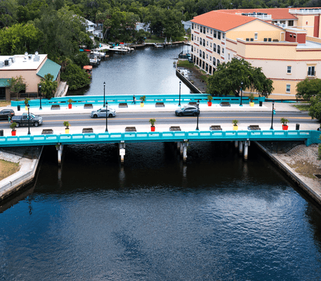 A teal bridge with cars and potted plants crosses over a river, with buildings and trees on either side of the water.