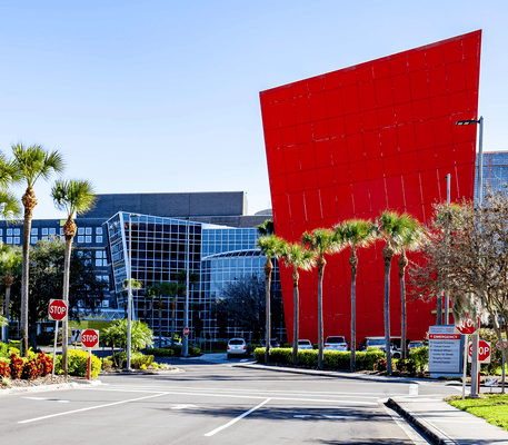Modern office building with a large, tilted red facade, glass windows, palm trees, and several stop signs at the entrance.