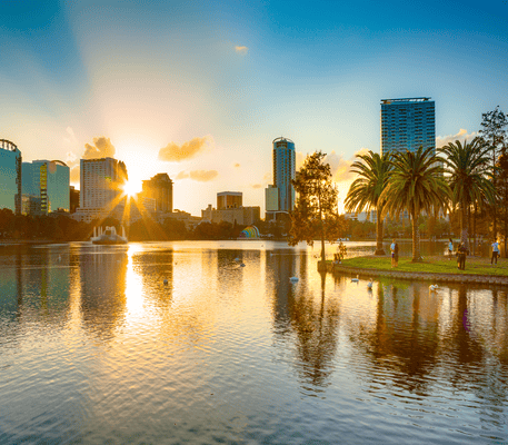 Sunset over a city skyline with high-rise buildings, palm trees, and people by a lake; sunlight reflects on the water’s surface.