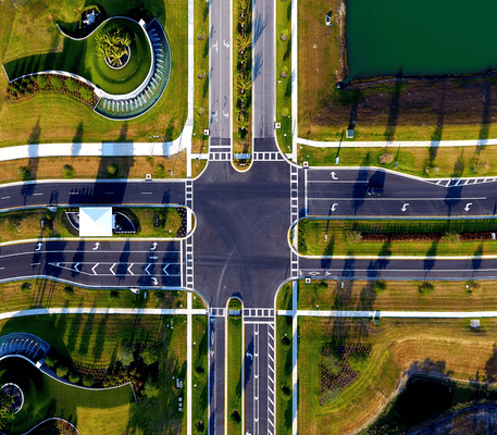 Aerial view of a large intersection with marked crosswalks, surrounding green lawns, landscaped plants, and nearby water features.