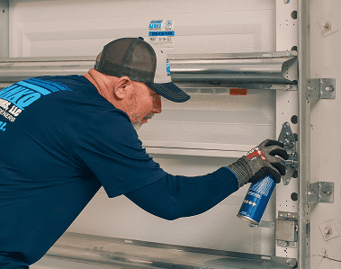 A man in work gloves and a cap sprays lubricant on a metal hinge of a garage door inside a garage.