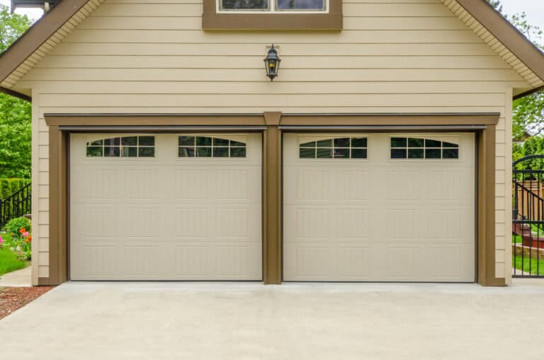 Two closed beige garage doors with small rectangular windows at the top, set in a tan house with brown trim and an outdoor light above the center.