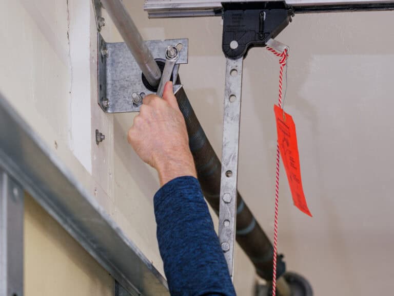 Person adjusting a metal rod with a wrench near a garage door spring mechanism; a red tag hangs nearby.