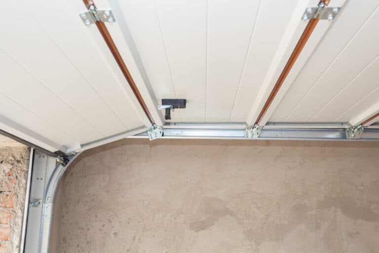 Partially opened white garage door showing metal tracks and torsion springs, with a beige unfinished wall in the background.