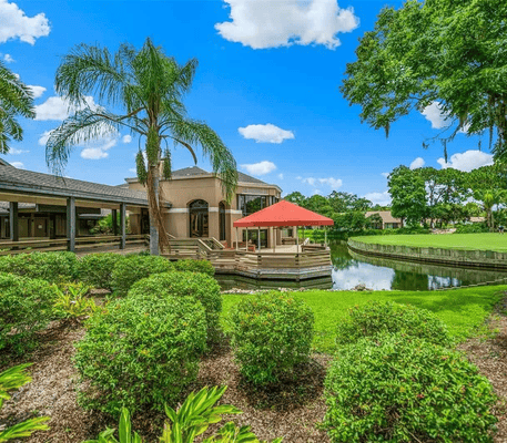Manicured green bushes and palm trees frame a building with a red umbrella by a pond, under a blue sky with scattered clouds.