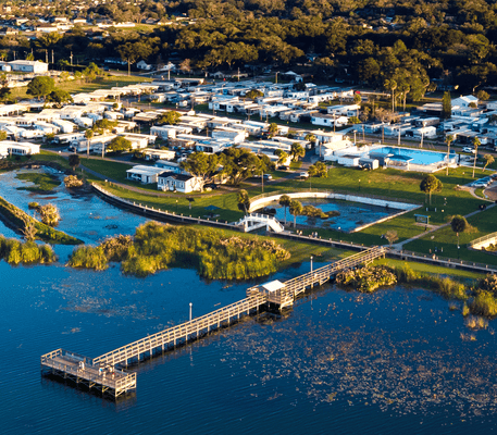 Aerial view of a lakeside residential area with a long wooden pier extending over the water, surrounded by greenery and nearby houses.