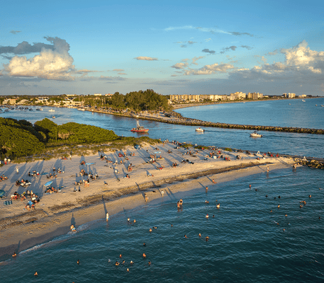 Aerial view of a crowded beach with people sunbathing and swimming, lush greenery, boats in the water, and buildings visible in the distance under a partly cloudy sky.