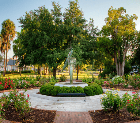 Circular garden with a central statue, surrounded by rose bushes, brick pathways, a bench, and large trees in a park setting under clear sky.